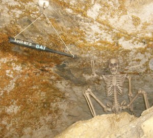 Skeleton sitting on rock in cave next to Tucker's Black Bat hanging from the ceiling