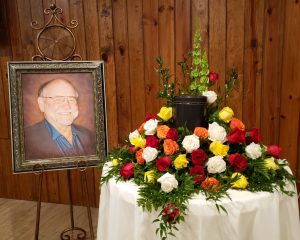 A picture of Ken next to a round table with multi-colored roses arranged around a round, black leather urn.