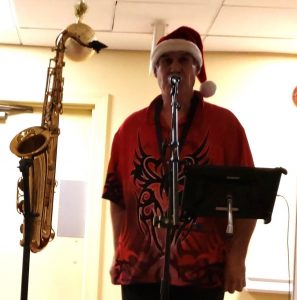Man standing behind a microphone wearing a Santa hat with a saxophone nearby